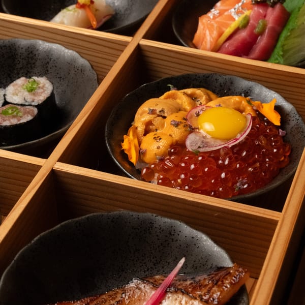 A wooden bento box filled with various Kase Sake & Sushi dishes, including uni, ikura, and tuna rolls.