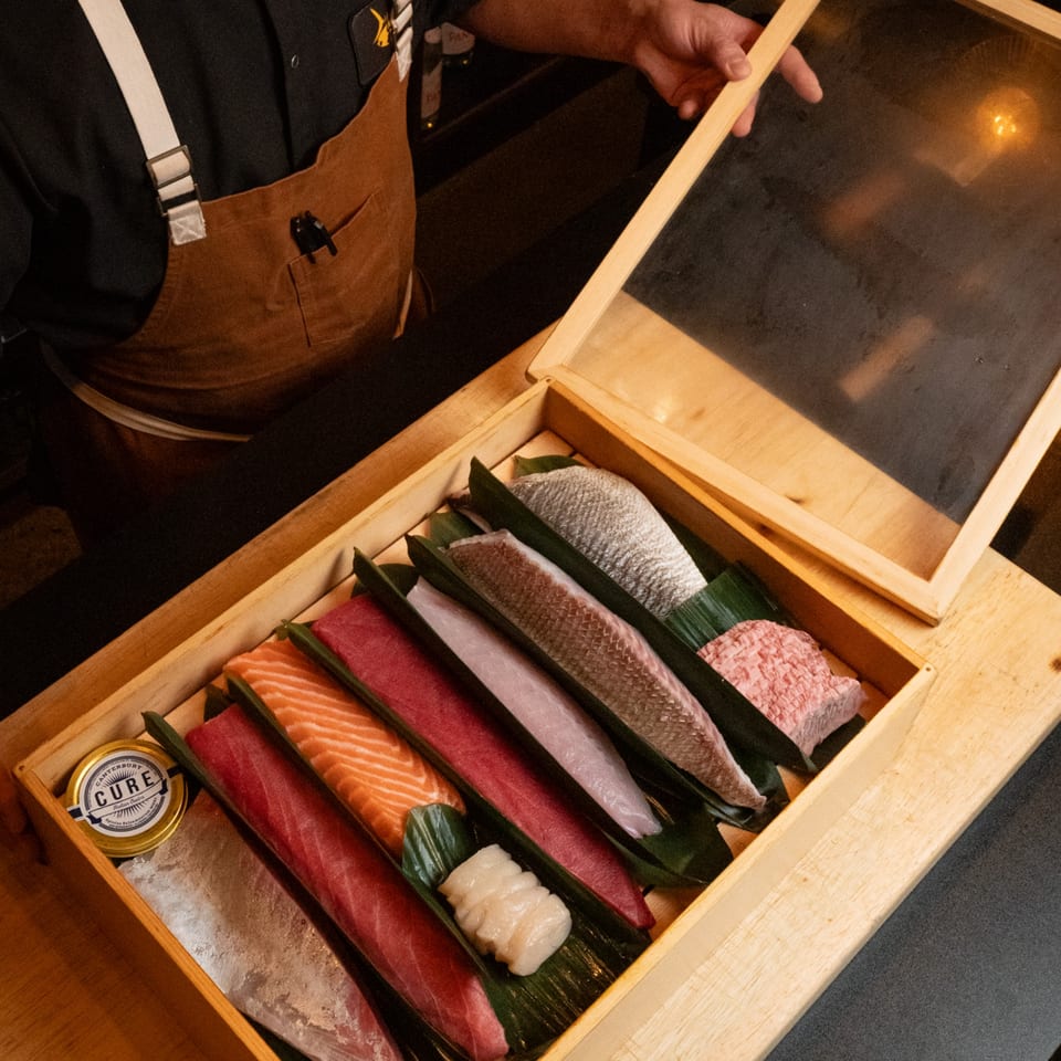 A chef opens a wooden box filled with various cuts of fresh fish and seafood for sushi.